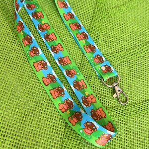 Könnte beinhalten: Ein blaues und grünes Lanyard mit einem sich wiederholenden Muster aus braunen Kühen, die Blumenkränze tragen. Das Lanyard hat einen silbernen Metallverschluss.