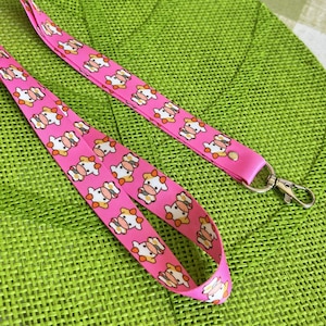 Könnte beinhalten: Rosafarbenes Lanyard mit einem sich wiederholenden Muster aus Comic-Kühen. Das Lanyard hat einen silbernen Metallverschluss.