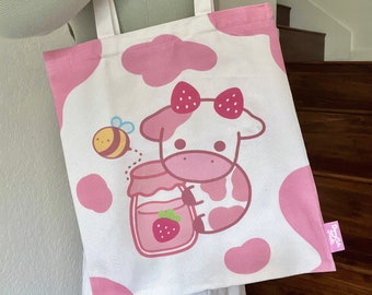 May include: A white tote bag with pink accents features a cartoon cow holding a jar of strawberry jam. A bee flies above the cow. The bag has a pink label that says "Dreamy Studio".
