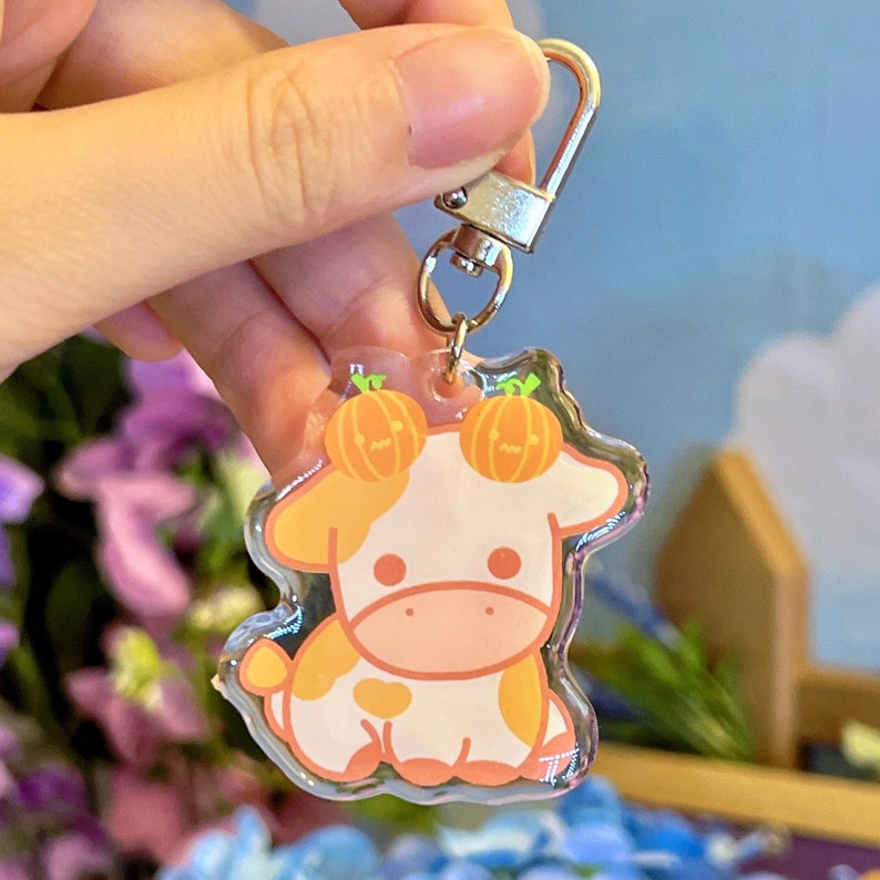 K&ouml;nnte beinhalten: Ein durchsichtiger Acryl-Schl&uuml;sselanh&auml;nger mit einer Comic-Kuh mit orangefarbenen K&uuml;rbis-Akzenten auf dem Kopf. Die Kuh ist wei&szlig; mit orangefarbenen Flecken und hat eine rosa Nase und Hufe.