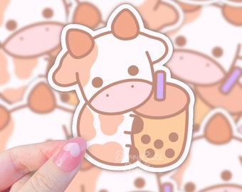 Puede incluir: Una pegatina con una vaca de dibujos animados con manchas marrones y blancas que sostiene una taza de té de boba con perlas de tapioca marrones. La vaca tiene la nariz y las mejillas rosas. La pegatina tiene un contorno blanco.