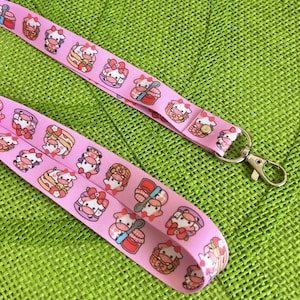 Könnte beinhalten: Rosafarbenes Lanyard mit einem sich wiederholenden Muster aus Comic-Kühen, die Hüte tragen und verschiedene Gegenstände wie Löffel, Milchkartons und Obstkörbe halten. Das Lanyard hat einen silbernen Metallschlüsselring.