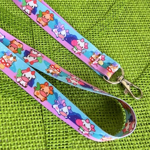 Könnte beinhalten: Ein blaues und rosa Lanyard mit einem sich wiederholenden Muster aus Comic-Kühen mit Flügeln und Blumen. Das Lanyard hat einen silbernen Metallverschluss.