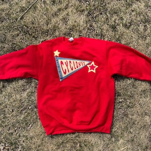May include: A red sweatshirt with a crew neck. The sweatshirt features a pennant design with the word "CYLONES" in red lettering, two yellow stars, and a blue denim-like patch.