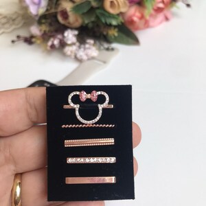 May include: A set of four rose gold rings with clear and pink rhinestones. The rings are displayed on a black velvet background. The top ring features a Mickey Mouse head with a pink bow.