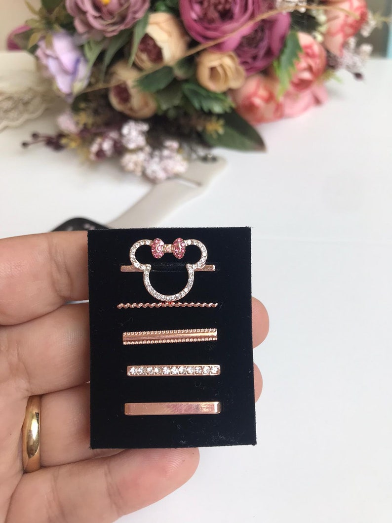 May include: A set of four rose gold rings with clear and pink rhinestones. The rings are displayed on a black velvet background. The top ring features a Mickey Mouse head with a pink bow.