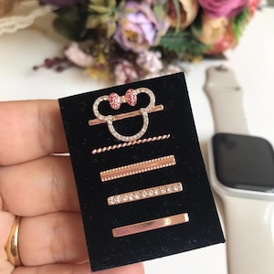 May include: A set of five rose gold hair clips with rhinestones. The clips feature a Mickey Mouse head design with a pink bow, a small chain, a straight bar with a beaded edge, a straight bar with rhinestones, and a plain straight bar.