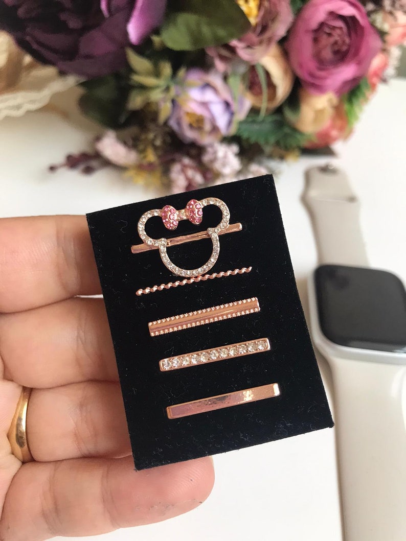 May include: A set of five rose gold hair clips with rhinestones. The clips feature a Mickey Mouse head design with a pink bow, a small chain, a straight bar with a beaded edge, a straight bar with rhinestones, and a plain straight bar.