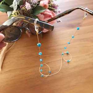 Könnte beinhalten: Eine Schildpatt-Sonnenbrille mit einer goldenen Brillenhalterungskette. Die Kette ist mit kleinen, runden, türkisfarbenen Perlen verziert. Im Hintergrund befindet sich ein Strauß aus rosa und lila Blumen auf einer Holzoberfläche.