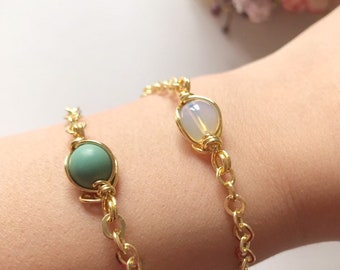 May include: Two gold chain bracelets with a green and a white oval stone charm. The bracelets are on a person's wrist.