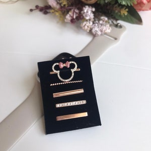 May include: A set of five gold-tone hair clips with rhinestones and a pink bow. The clips are displayed on a black velvet background.