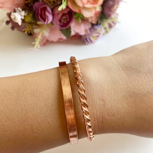Puede incluir: Dos pulseras de cobre se muestran en una muñeca. Una es una banda plana y simple, y la otra tiene un diseño de cuerda trenzada. Las pulseras son de un color marrón rojizo cálido, y el fondo presenta un ramo de flores rosas y moradas.