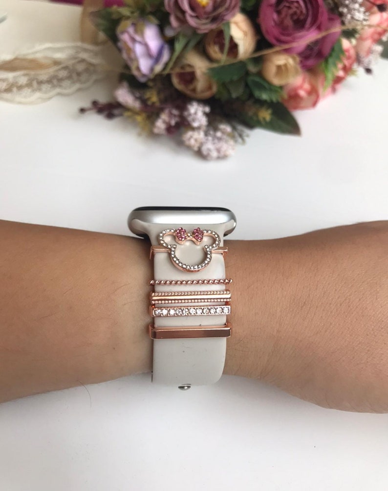 May include: A white silicone Apple Watch band with a rose gold Mickey Mouse charm and four rose gold bands with clear rhinestones.