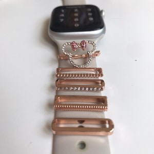 May include: A white Apple Watch band with a Mickey Mouse charm and several rose gold watch band charms with clear rhinestones.
