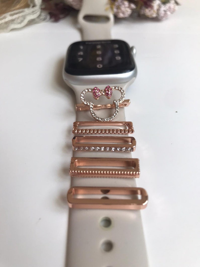 May include: A white Apple Watch band with a Mickey Mouse charm and several rose gold watch band charms with clear rhinestones.