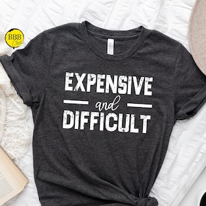 May include: A dark gray t-shirt with the text "EXPENSIVE and DIFFICULT" printed in white.