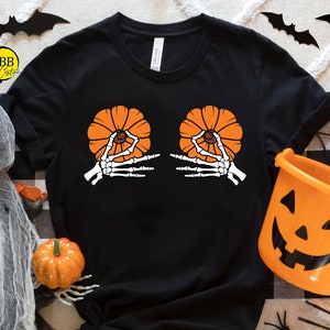 May include: Black t-shirt with a Halloween-themed design. The graphic features two orange pumpkins held by white skeleton hands. The shirt is surrounded by Halloween decorations, including pumpkins, spiders, and bats.