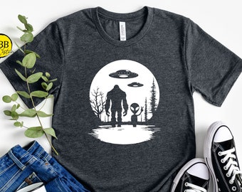 Könnte beinhalten: Ein dunkelgraues T-Shirt mit einem weißen Aufdruck eines Bigfoots und eines Außerirdischen, die zusammen vor einem Vollmond stehen. Der Aufdruck enthält auch ein UFO über dem Mond.