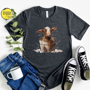 May include: A dark gray t-shirt with a graphic of a brown baby goat sitting in a bucket. The goat is looking at the camera and has a white spot on its forehead. The bucket is brown and has a white rim. The t-shirt is on a white background.