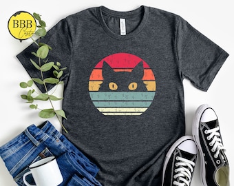 May include: A dark gray t-shirt with a retro sunset design featuring a black cat silhouette with yellow eyes.