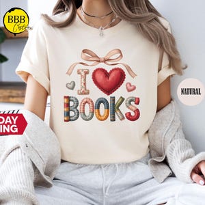 May include: Cream-colored t-shirt with the phrase "I love books" in a decorative design. The design includes a red heart, a bow, and the word "BOOKS" made to look like stacked books. The shirt has a round neckline and short sleeves.