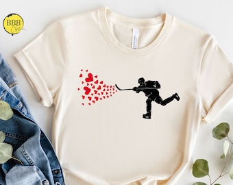 May include: A cream-colored t-shirt with a black silhouette of a hockey player blowing hearts. The player is holding a hockey stick and is skating towards the right. The hearts are red.