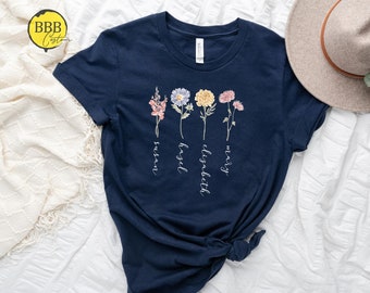 Può includere: Una maglietta blu navy con un design floreale e i nomi "Mary", "Elizabeth", "Hazel" e "Susan" scritti verticalmente sotto i fiori.