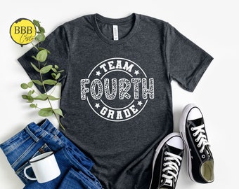 Könnte beinhalten: Ein dunkelgraues T-Shirt mit einem weißen Kreisdesign, auf dem in weißer Leopardenmuster-Schrift "Team Fourth Grade" steht. Das Shirt liegt flach auf einer weißen Oberfläche mit einem Paar schwarzen Turnschuhen und einer blauen Jeans.