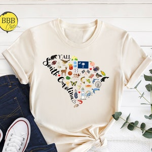 May include: A beige t-shirt with a heart-shaped design featuring various icons representing South Carolina. The text "Y'all South Carolina" is written inside the heart.