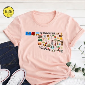 May include: A pink t-shirt with a graphic design of the state of Oklahoma. The design includes various icons representing the state, such as a bison, a windmill, and a cowboy boot. The text "The Sooner State" and "Oklahoma" are also included in the design.
