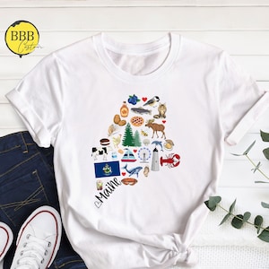May include: White t-shirt with a heart-shaped design featuring various icons representing Maine, including a lobster, lighthouse, pine tree, and the state flag. The text "Maine" is written below the design.