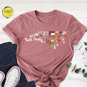 May include: A pink t-shirt with a graphic of the state of North Carolina, featuring various icons representing the state, including mountains, a lighthouse, a pig, a peach, a bee, a football, and the state flag. The text "Y'all" and "North Carolina" are also included in the graphic.