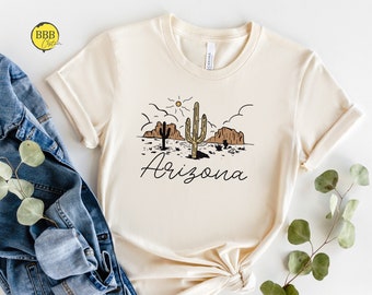 May include: A beige t-shirt with a graphic of a cactus, mountains, and a sun in a desert landscape. The text "Arizona" is printed below the graphic.