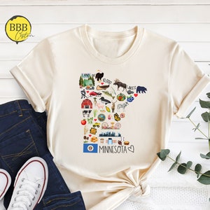 以下が含まれることがあります： ミネソタ州のグラフィックが描かれたベージュのTシャツ。グラフィックは、湖、ヘラジカ、クマ、農場、都市のスカイラインなど、州に関連するさまざまなアイコンで構成されています。「Land of 10,000 Lakes」と「Minnesota」のテキストがグラフィックに含まれています。