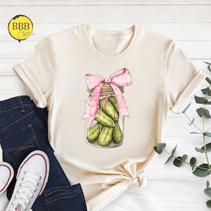 May include: A beige t-shirt with a graphic of a mason jar filled with pickles and tied with a pink ribbon.