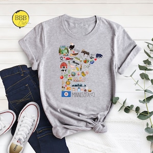 以下が含まれることがあります： ミネソタ州のグラフィックが描かれたグレーのTシャツ。グラフィックには、湖、木、ヘラジカなど、州に関連するものを表すさまざまなアイコンが含まれています。「Land of 10,000 Lakes」と「Minnesota」のテキストもグラフィックに含まれています。