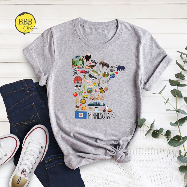 以下が含まれることがあります： ミネソタ州のグラフィックが描かれたグレーのTシャツ。グラフィックには、湖、木、ヘラジカなど、州に関連するものを表すさまざまなアイコンが含まれています。「Land of 10,000 Lakes」と「Minnesota」のテキストもグラフィックに含まれています。
