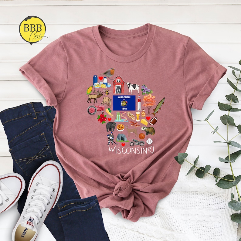 May include: A mauve t-shirt with a heart-shaped design featuring various icons representing Wisconsin. The icons include a cow, a cheese wedge, a beer stein, a fish, a football, and a red barn. The text "WISCONSIN" is written below the heart.