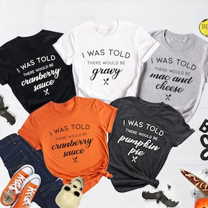 Op de afbeelding: Vijf verschillende T-shirts met een Thanksgiving-thema. Elk shirt heeft een ander voedsel erop, waaronder cranberry-saus, jus, macaroni en kaas, pompoentaart. De shirts zijn allemaal wit, zwart, grijs, oranje en donkergrijs.