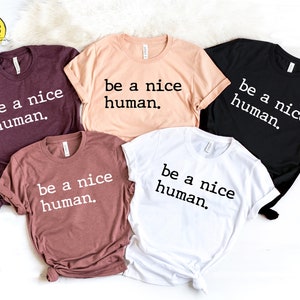 May include: Five t-shirts in various colors, including burgundy, peach, mauve, black, and white. Each shirt features the phrase "be a nice human" in a bold, sans-serif font. The shirts are neatly arranged, showcasing the design.