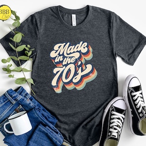 Op de afbeelding: Een donkergrijs T-shirt met een retro-stijl grafische afbeelding met de tekst "Made in the 70's". De afbeelding is in regenboogkleuren met een vintage verweerde look.