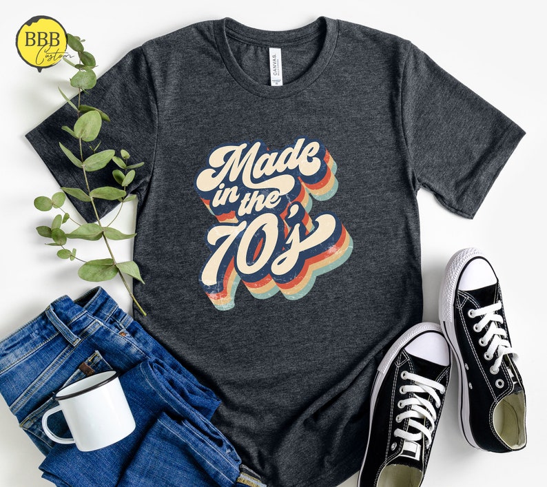 Op de afbeelding: Een donkergrijs T-shirt met een retro-stijl grafische afbeelding met de tekst "Made in the 70's". De afbeelding is in regenboogkleuren met een vintage verweerde look.
