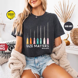 May include: Dark gray t-shirt with a graphic of syringes in various sizes and colored tips. The text "SIZE MATTERS" is below the syringes, with "#nurselife" underneath. The shirt is worn by a person.