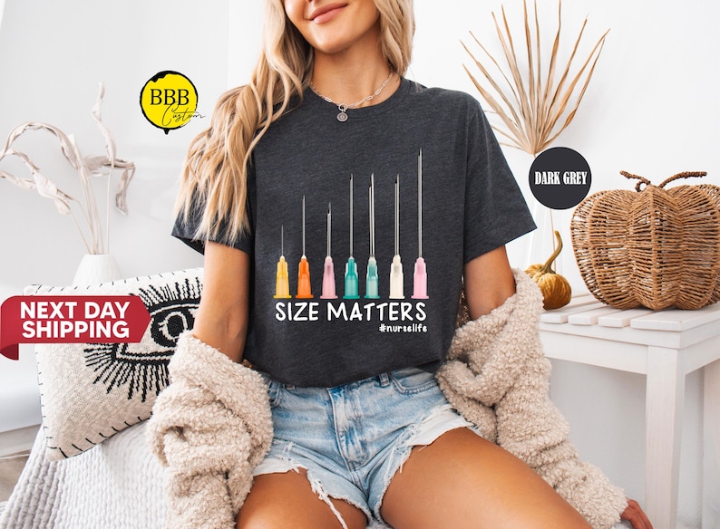 May include: Dark gray t-shirt with a graphic of syringes in various sizes and colored tips. The text "SIZE MATTERS" is below the syringes, with "#nurselife" underneath. The shirt is worn by a person.