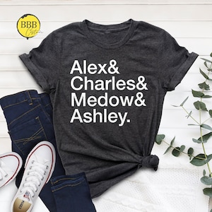 May include: Dark gray t-shirt with the names Alex, Charles, Medow, and Ashley printed in white. The shirt is displayed with blue jeans and white canvas sneakers. The shirt has a crew neck and short sleeves.