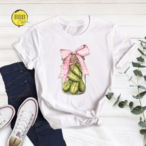 May include: White t-shirt with a graphic of a glass jar filled with green pickles and tied with a pink ribbon.