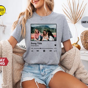 May include: Light grey t-shirt featuring a graphic of a music player interface with a photo of three women. The text on the graphic reads "Song Title" and "Artist Name". The shirt is worn by a person wearing denim shorts.