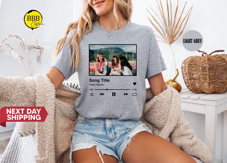 May include: Light grey t-shirt featuring a graphic of a music player interface with a photo of three women. The text on the graphic reads "Song Title" and "Artist Name". The shirt is worn by a person wearing denim shorts.