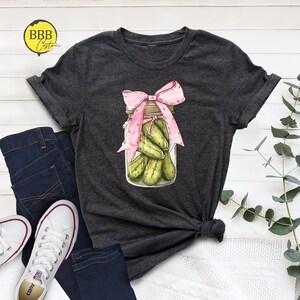May include: A dark gray t-shirt with a graphic of a glass jar filled with green pickles and topped with a pink bow.
