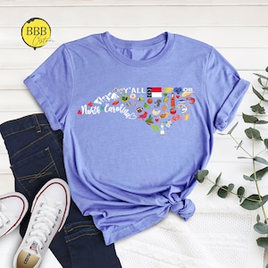 May include: A light blue t-shirt with a graphic design of a heart-shaped outline of North Carolina. The graphic includes various icons representing the state, including a lighthouse, a beach umbrella, a race car, a football, a chicken, a peach, and the state flag. The text "Y'all" and "North Carolina" are included in the graphic.